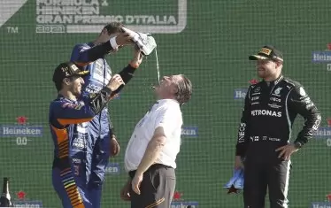 Победителят в "Гран при" на Италия Даниъл Рикардо (вляво) окуражава съотборника си Ландо Норис да налива шампанско от обувката на австралиеца в устата на изп. директор на "Макларън" Зак Браун пред смаяния поглед на Валтери Ботас от "Мерцедес" (вдясно).