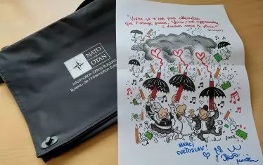 Нещо смешно от България: журналистическа чантичка с надпис НАТО, комбинирана с усмихната карикатура от Плантю.